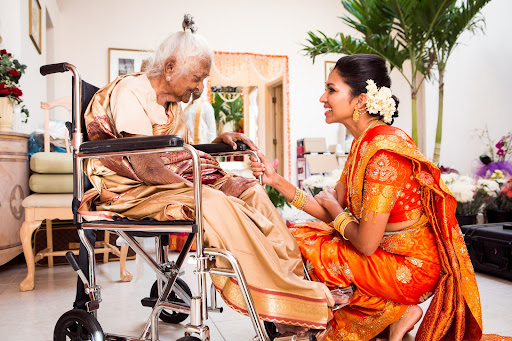 A young bride in a red Indian wedding gown leans in to her grandmother in a wheelchair