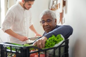 Older man sitting with a box of food, younger man in background